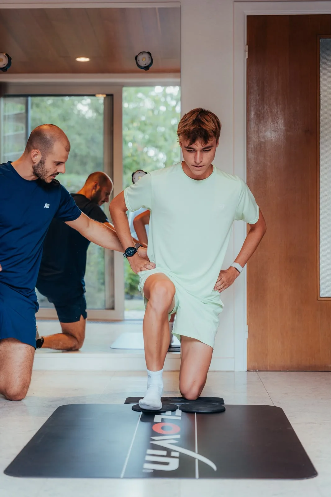 Kinésithérapeute accompagnant un jeune patient dans un exercice de fente sur tapis.